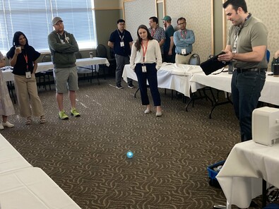 An adult male with a tablet codes a Sphero robot in a room while adults stand and view.