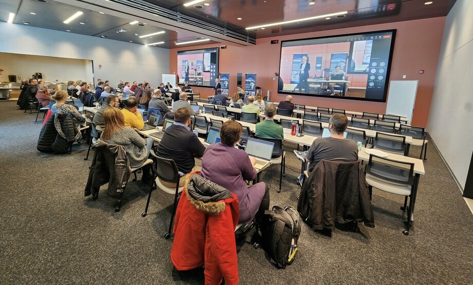 Panoramic image of a conference room with two display screens. Adults sit at rectangular tables while the screens display individuals joining via TEAMS